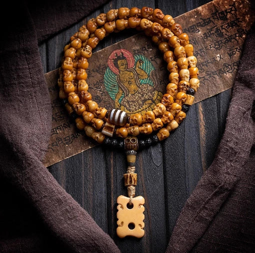 A handcrafted Tibetan yak bone 108 mala necklace with small carved skulls, arranged over a vintage canvas painting of a seated figure in a traditional Himalayan style on a dark wooden background framed by brown textured cloth.