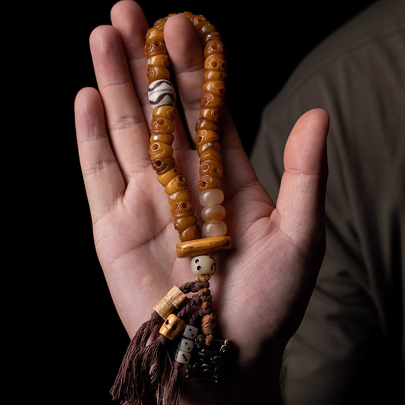 Tibetan Ox Bone Skull Bracelet—Energy A hand holding a handcrafted Tibetan yak bone bracelet with aged brown beads, a patterned focal bead, and a dark brown decorative tassel against a dark background.